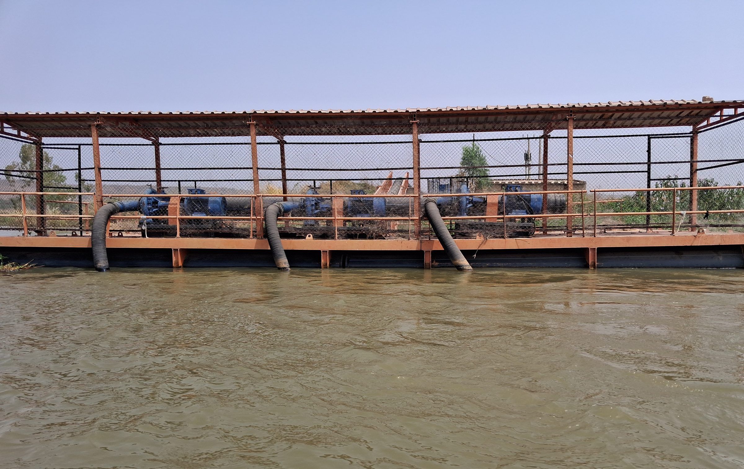 Structure métallique couverte posée sur l'eau, abritant une rangée de pompes reliées à des conduites plongeant dans le fleuve.